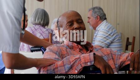 Leaning back senior man in wheelchair, staff in scrubs placing hand on shoulder in common room Stock Photo