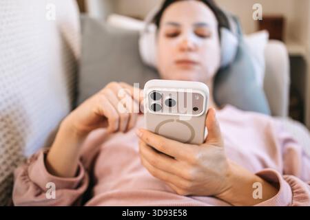 Female individual wearing headphones is comfortably lying on a couch, engaging with smartphone, surrounded by soft cushions, creating a relaxed atmosp Stock Photo