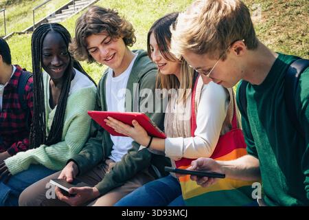 Group of multicultural teens sitting on steps outside, smiling and using digital devices together, enjoying social interaction. Stock Photo