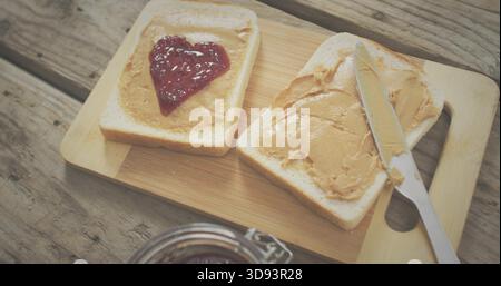 Showing two bread slices with peanut butter on bamboo board at kitchen table, heart-shaped jam Stock Photo