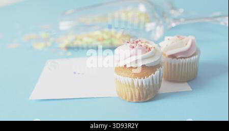 Showing two cupcakes with pink swirl frosting, sprinkles on pale-blue table, white card, copy space Stock Photo
