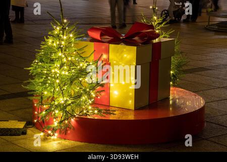 A large golden gift box with a red ribbon sits among small decorated Christmas trees with glowing fairy lights Stock Photo