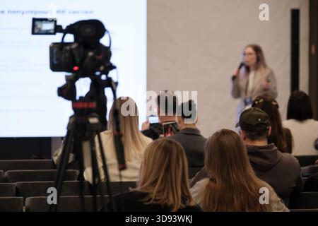Speaker with microphone presents to attentive audience while camera captures session, large projection screen displays slides; audience seated in rows; professional event atmosphere,  Stock Photo