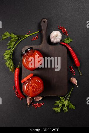 Homemade chili sauce jars on a dark cutting board, surrounded by fresh red chilies, garlic, parsley and scattered pink peppercorns for a vibrant, spic Stock Photo