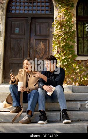 Two friends sit on stone steps outside a historic building, laughing and sharing a moment. One holds a coffee cup, while the other displays something Stock Photo