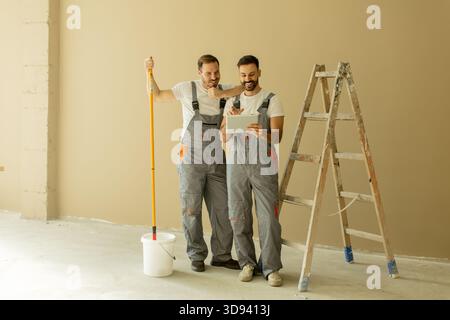 Two men wearing overalls smile while looking at a tablet in a room with light brown walls. A step ladder and paint bucket are next to them as they dis Stock Photo