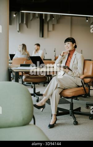 A woman dressed in a light suit sits confidently in a coworking space. She holds a tablet and smiles while engaging with coworkers. The setting is mod Stock Photo