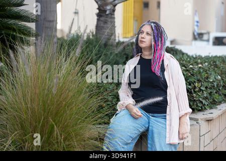 Portrait of a middle aged woman with colorful dreadlocks posing outdoors next to tall ornamental grass. Stock Photo