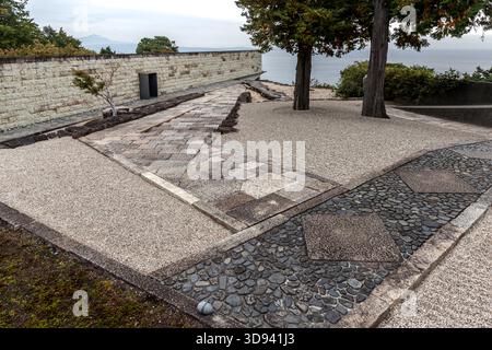 A paved outdoor area featuring a mosaic of various stone materials – granite, river pebbles and concrete slabs – alongside a rubble-stone wall overlooking Sagami Bay in Odawara, Kanagawa Prefecture, Japan. The silhouette of a mountain can be made out in the haze on the horizon; on a clear day, Mount Fuji (3,776 m) is visible from Odawara Stock Photo