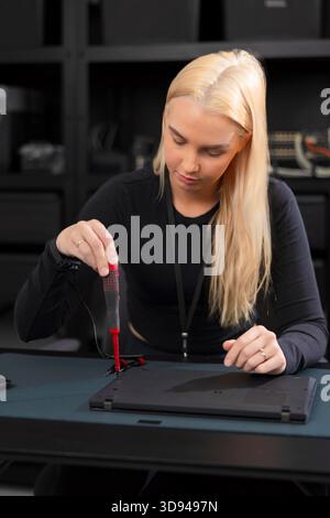 Young Caucasian woman with blonde hair repairs laptop using a screwdriver in a tech workshop Stock Photo