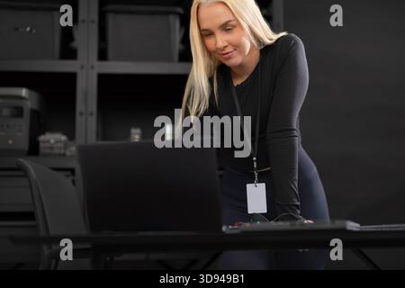 Female IT technician with laptop examining work desk in a tech office setting Stock Photo