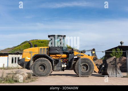 Bright yellow wheel loader stands behind metal fence on dusty construction ground. Sunlight highlights large tires, cabin and bucket while green hill Stock Photo