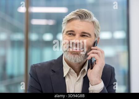 Mature businessman having a cheerful conversation on his mobile phone in an office setting, representing successful communication, professionalism, and positive business interactions Stock Photo