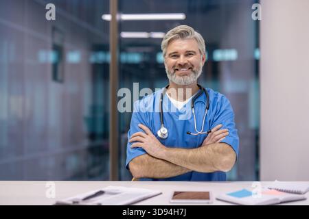 Healthcare professional wearing blue scrubs and stethoscope, sitting at a desk with arms crossed, smiling at the camera in a modern hospital or clinic setting Stock Photo