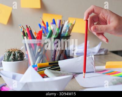 hand holding a red colored pencil ready to draw set against a busy desk with a cactus, pen holders and paper crafts - creativity, planning, education Stock Photo