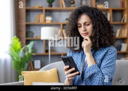 Young woman with curly hair sitting on a couch at home, focusing on a smartphone screen with a concerned expression, contemplating a difficult message or shocking news Stock Photo