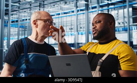 Photovoltaics smart factory team leader teaching intern how to fix damaged IoT sensors using laptop. Industry 4.0 solar panels plant experienced engineer guiding trainee in reducing downtime, camera A Stock Photo