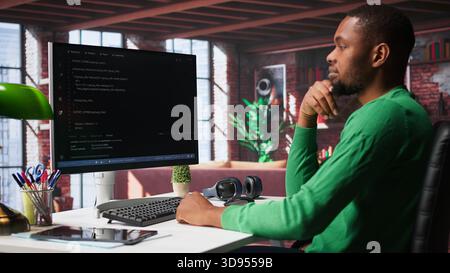 Black technician in home office looks at UI on computer monitor, typing and executing programming code. IT worker using coding application to write and test code in modern apartment. Camera B. Stock Photo
