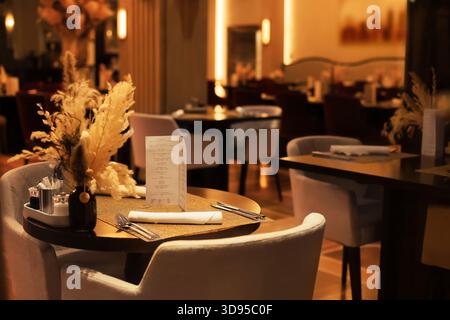 Elegant restaurant table setting with menu, cutlery and dried flowers in vase under warm ambient lighting. High quality photo Stock Photo