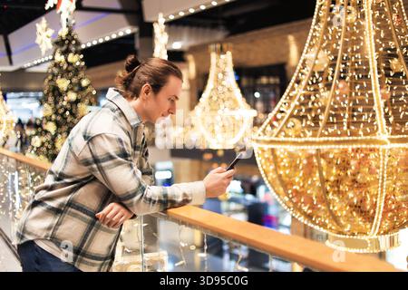 Young man leaning on railing using phone amidst festive Christmas lights and decorations in a shopping mall. High quality photo Stock Photo
