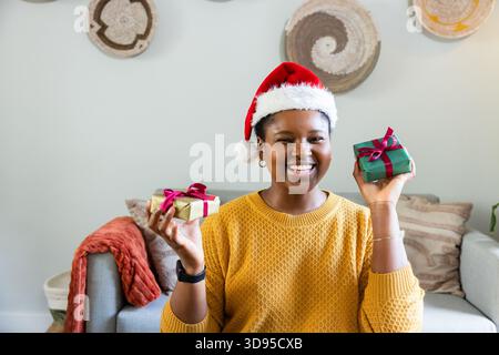 African American woman sitting in living room wearing Santa hat and sweater holding two gift boxes Stock Photo