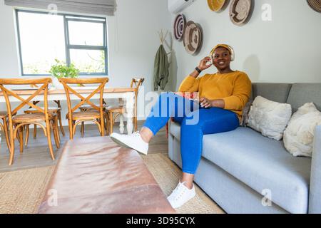 Adult African American woman wearing sweater sitting on sofa wearing headphones at home, copy space Stock Photo