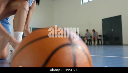 Resting basketball occupying center on blue court, kneeling player tying shoe in light blue uniform Stock Photo