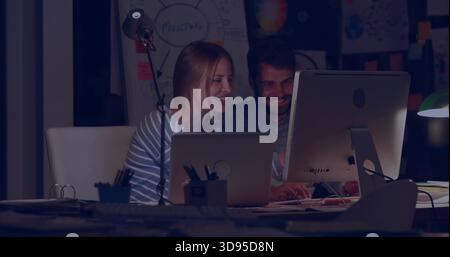 Leaning, pair wearing dark shirt, striped top reviewing code in dim workspace with monitor, laptop Stock Photo