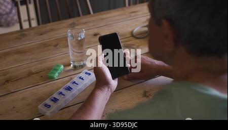Holding smartphone, senior man in green shirt checking pill organizer on wooden table, copy space Stock Photo