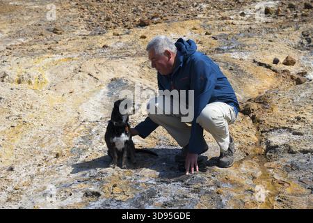 Man playing with dog in a rocky, natural environment, dressed in outdoor clothing, dog and man looking at each other Stock Photo