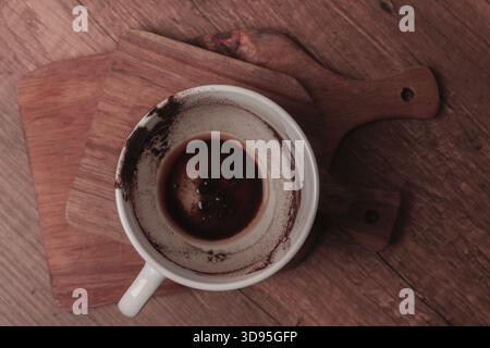 A warm coffee cup sits on a wooden cutting board, showing traces of dark liquid and coffee grounds. This scene captures a moment of relaxation and com Stock Photo