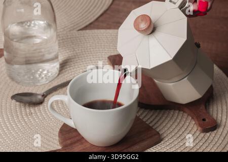 Freshly brewed coffee is being poured from a stovetop espresso maker into a white cup on a wooden table. A clear glass water bottle sits nearby, addin Stock Photo