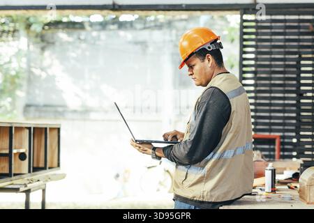 Male carpenter holding a laptop in the carpentry workshop. Cabinetmaker using a laptop in the carpentry workshop Stock Photo