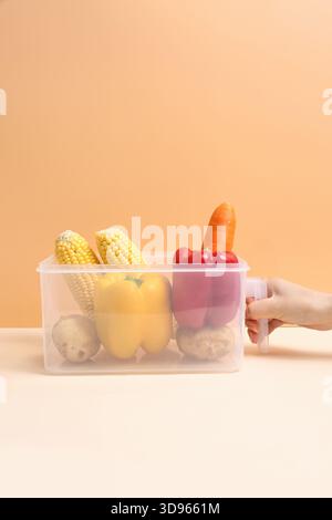 Hand holding clear plastic food storage container with red peppers and potatoes on beige background Stock Photo