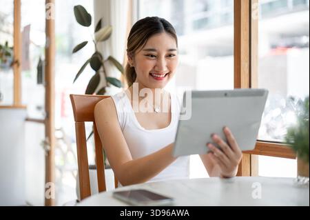 Pretty asian woman holding and looking at the tablet while sitting at round table beside glass wall in cafe. Free Time Relaxing, Chilling Outside. Stock Photo