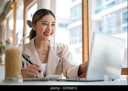 Smart asian woman holding pen over notebook looking and touching on laptop as sitting with coffee at cafe's table. Working or Study, Chilling Outside. Stock Photo