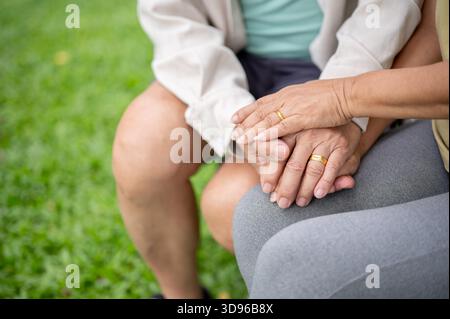 Close up of old man and woman's hands with gold wedding rings clasping together while sitting in park. Elderly couple, Green Nature, Chilling Outdoor. Stock Photo