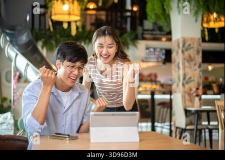 Standing asian woman and sitting man looking happy excited at tablet on wood table in cafe or canteen. Working or study, Assignment or Group project. Stock Photo