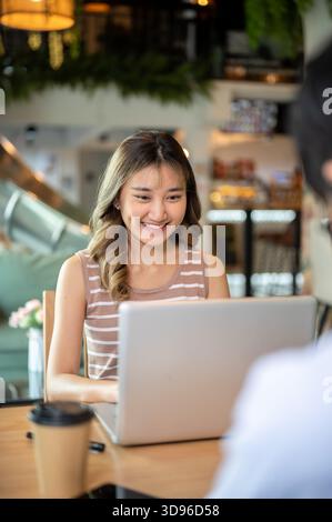 Asian woman using or looking at laptop while sitting with a man at wooden table in cafe or canteen. Working or study, Assignment or Group project. Stock Photo