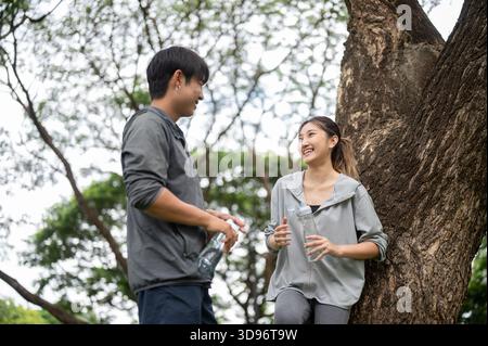 Asian man holding water bottle smiling talking with woman in sportswear leaning against a tree in park. Young Couple, Training Tracksuit. Green Nature Stock Photo