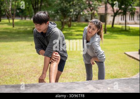 Asian man and a woman in sportswear stretching their arm and leg on cement stair in park's courtyard. Young Couple, Training Tracksuit. Green Nature. Stock Photo