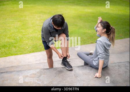 Asian man in sportswear adjusting a sock while a woman sitting on cement bench in park's courtyard. Young Couple, Training Tracksuit. Green Nature. Stock Photo