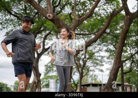 Asian man and a woman wearing sportswear laughing during a running or jogging on pavement in park. Young Couple, Training Tracksuit. Green Nature. Stock Photo