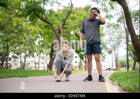 Asian woman in sportswear sits tying shoelace and a man wiping sweat after running on pavement in park. Young Couple, Training Tracksuit. Green Nature Stock Photo