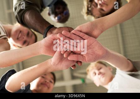 Diverse group of teenagers stacking hands in unity, viewed from below, including Caucasian, Black, and Asian individuals, showing teamwork and cooperation in collaborative setting Stock Photo