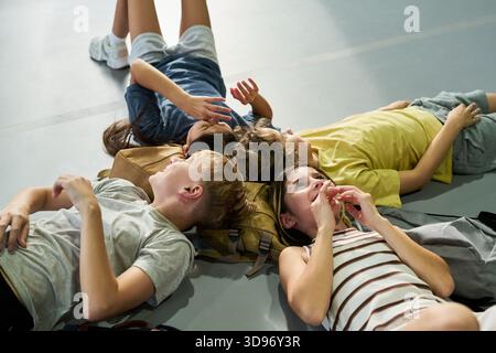 Group of four teenagers lying on floor, relaxing together and talking, two Caucasian boys and two Caucasian girls with backpacks visible, engaging in casual conversation indoors Stock Photo