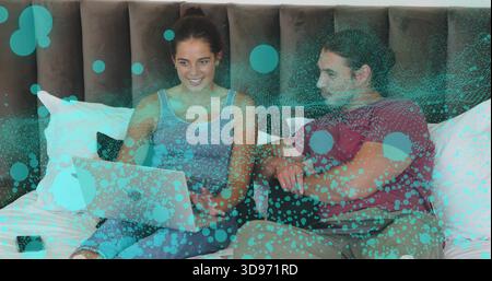 Sitting woman using laptop while man leaning in on bed, casual clothes, teal-green overlay Stock Photo