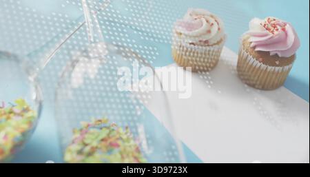 Showing two sprinkled cupcakes on pastel-blue table, inverted glasses, dotted light, copy space Stock Photo