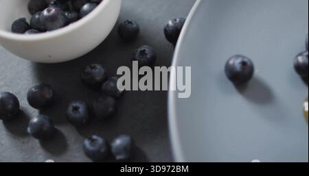 Glistening fresh blueberries resting on dark matte tabletop, with white bowl and gray plate Stock Photo