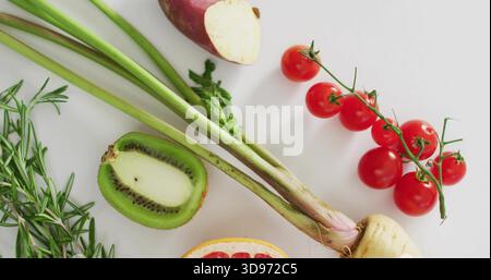 Composing produce flat-lay on white table, showing cherry tomatoes vine, halved kiwi, rosemary Stock Photo